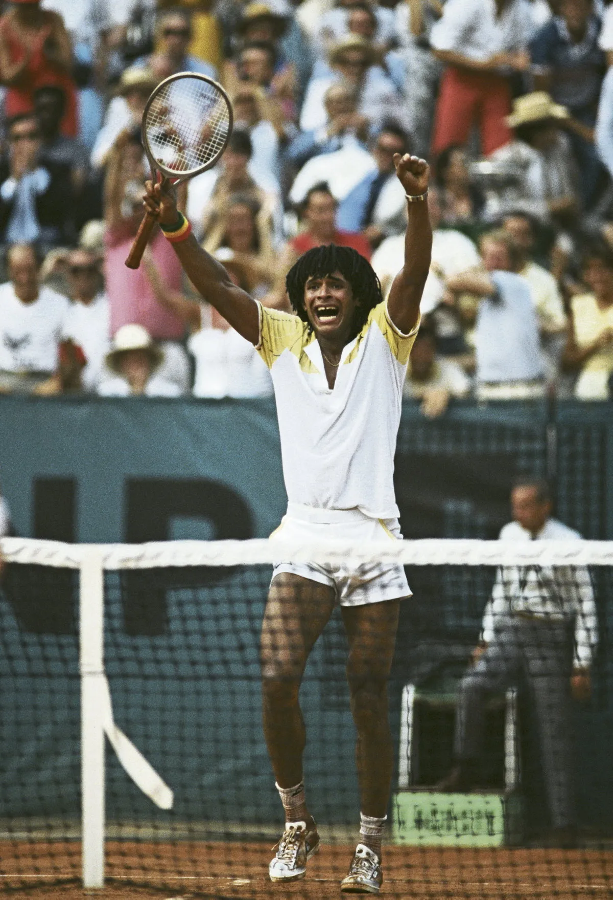Yannick Noah, vêtu de blanc, lève les bras vers le ciel, raquette dans la main droite, pour célébrer sa victoire à Roland Garros.